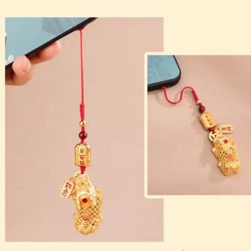 Gold-plated Pixiu phone charm with red braided lanyard attached to a smartphone case.
