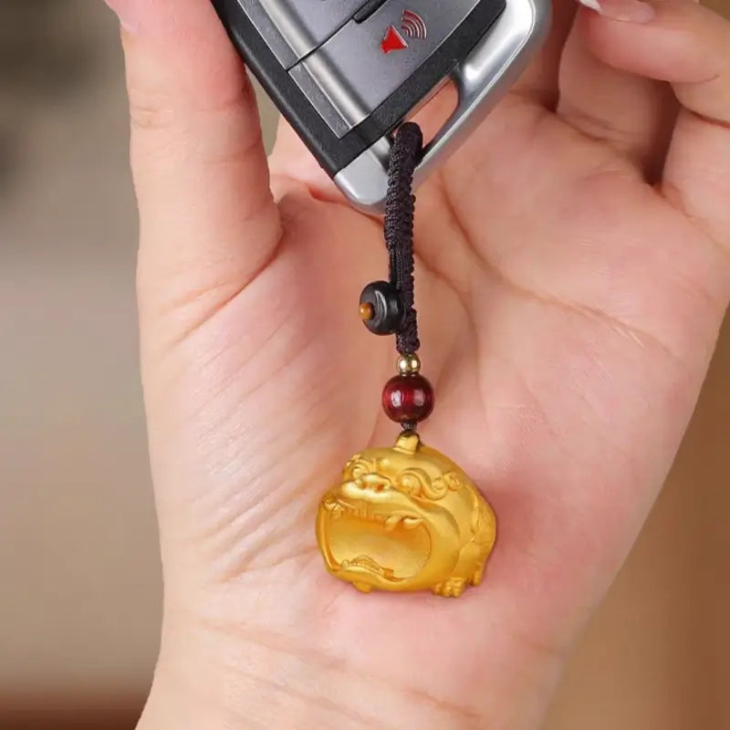 Hand holding a 24K gold-plated brass Pixiu keychain pendant attached to a car key remote.