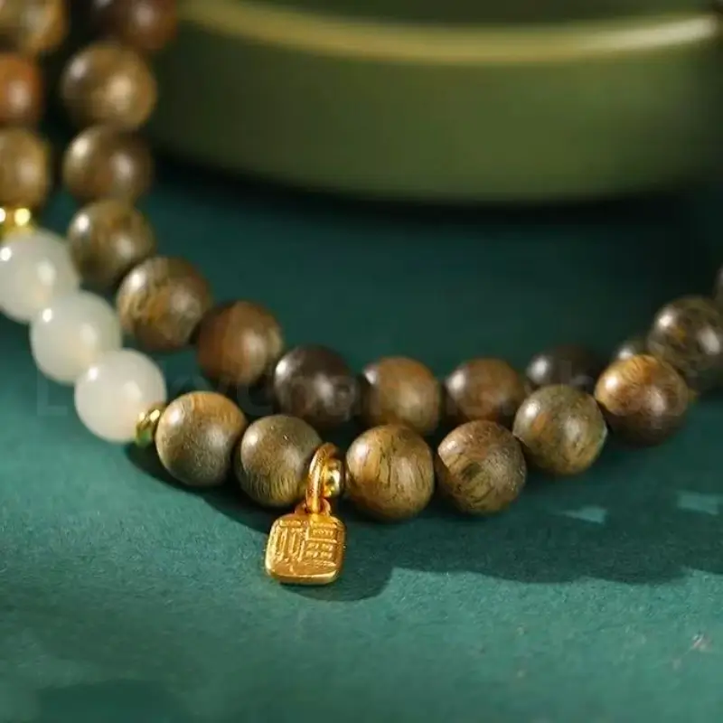 Extreme close-up of a 6mm natural green sandalwood bracelet showcasing a gold-plated fortune charm and glossy white jade beads on a green surface.