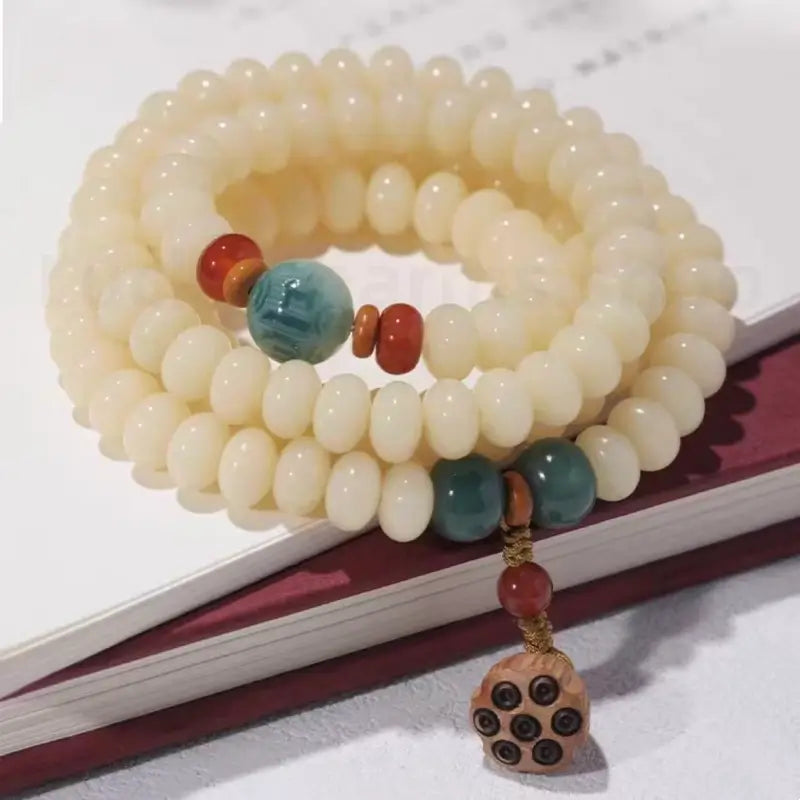A triple-strand white jade bodhi seed prayer bracelet stacked neatly on top of a book, featuring a dangling 14mm wooden lotus seedpod.