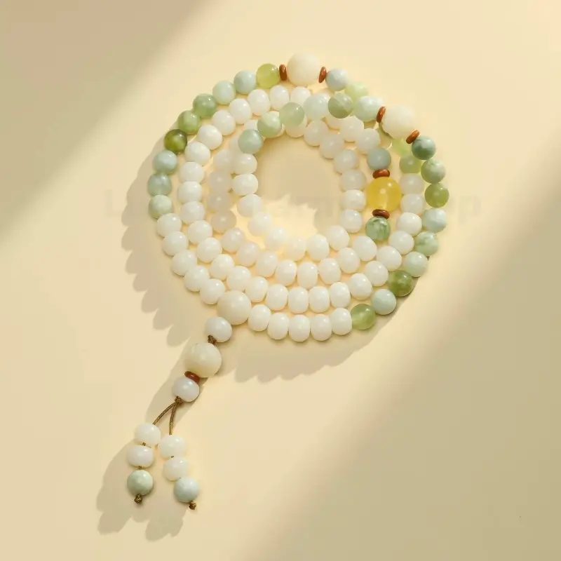 Coiled 108 white jade Bodhi seed mala bracelet with green jade spacers and tassel on beige background.