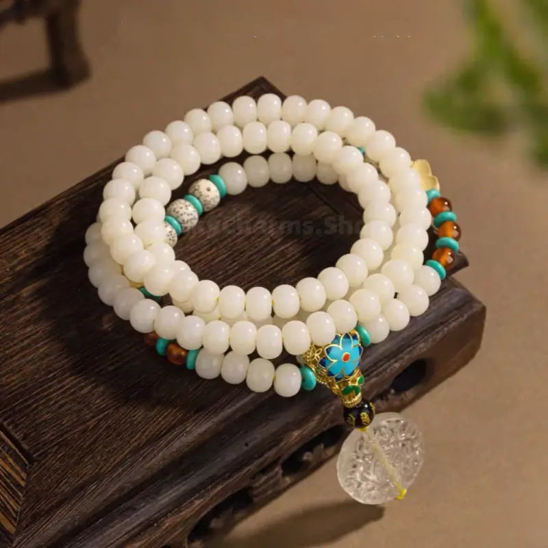 108 white jade Bodhi seed mala bracelet with blue floral bead and carved clear crystal pendant on dark wood stand.
