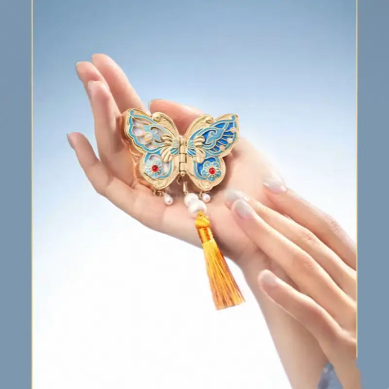 Blue enamel butterfly solid perfume compact with yellow tassel displayed in open hand.