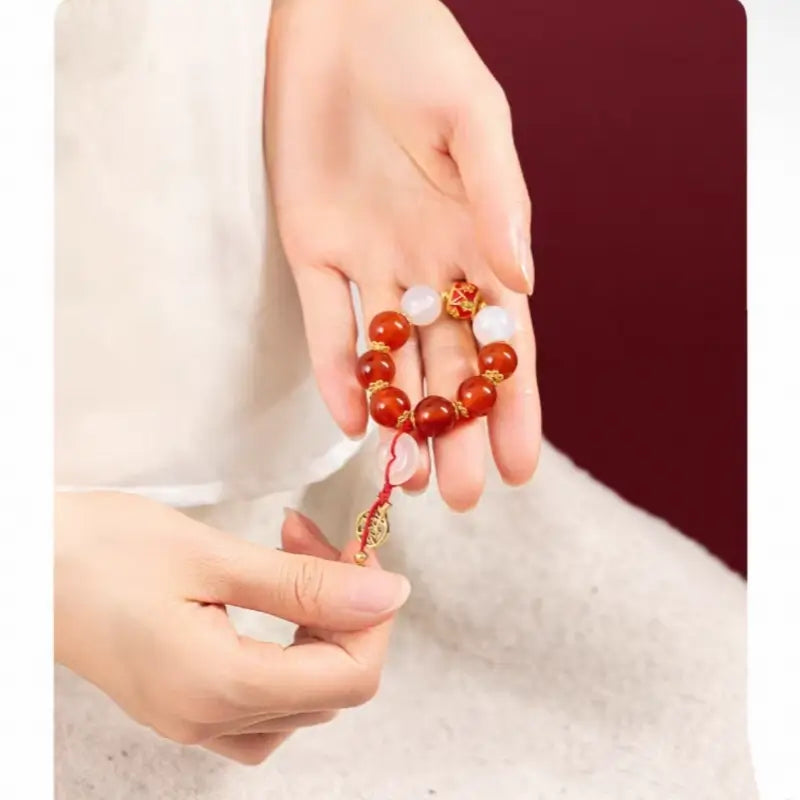 Hand holding red agate beaded phone strap with white quartz ring and gold fortune coin.