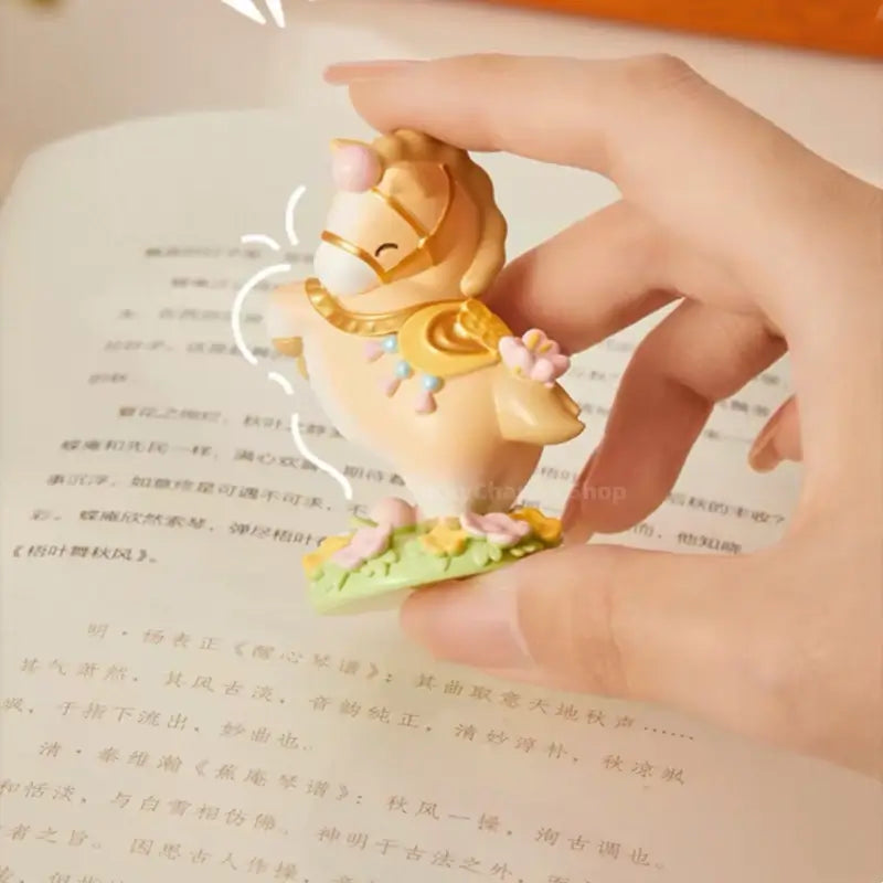 Hand holding miniature beige Palace Museum fortune horse figurine above an open book.