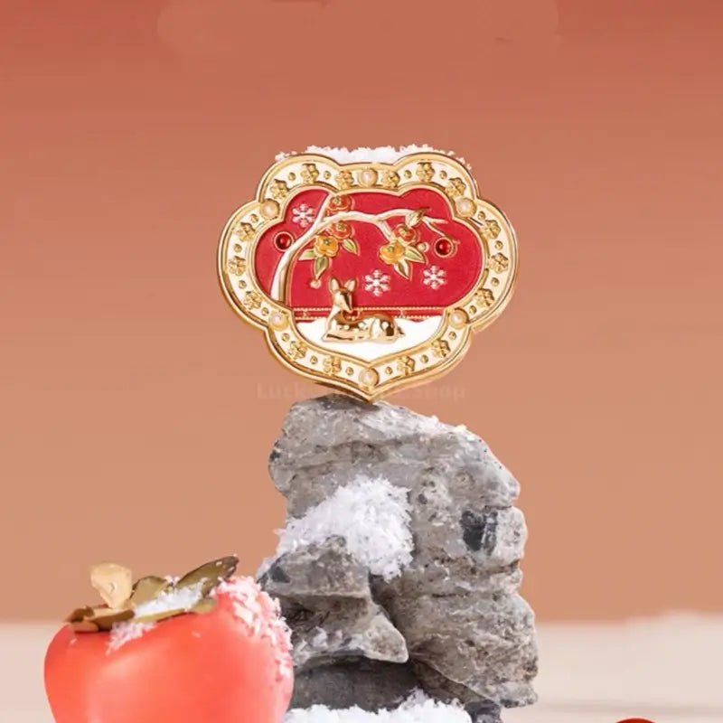Palace Museum red enamel Ruyi solid perfume compact with deer motif resting on snow-covered rock next to persimmon.