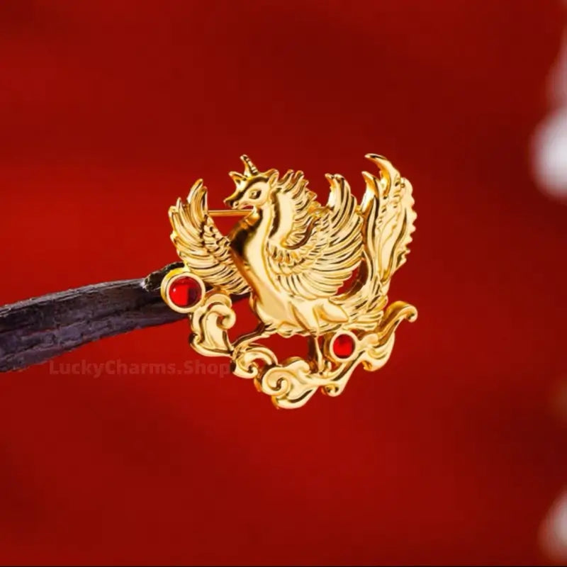 Gold-plated phoenix brooch with red gem accents resting on dark wood display stand.