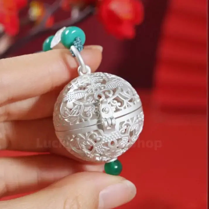 Hand holding a sterling silver filigree sphere sachet pendant with green jade bead accents.