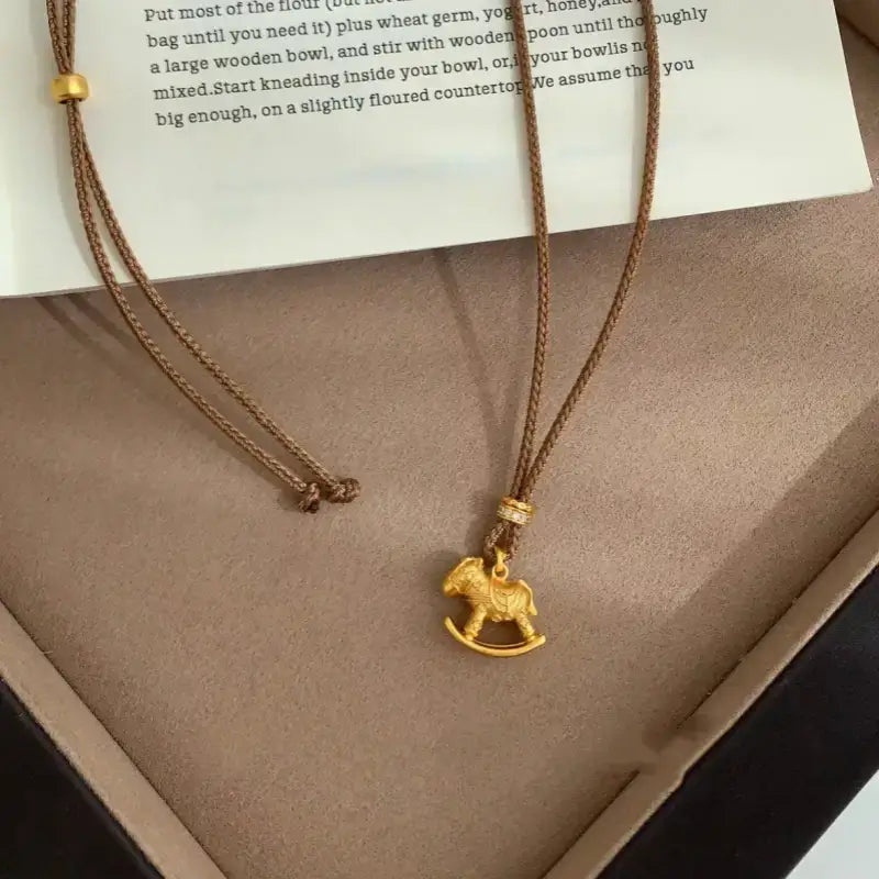 Gold rocking horse pendant necklace with brown woven cord next to a book.