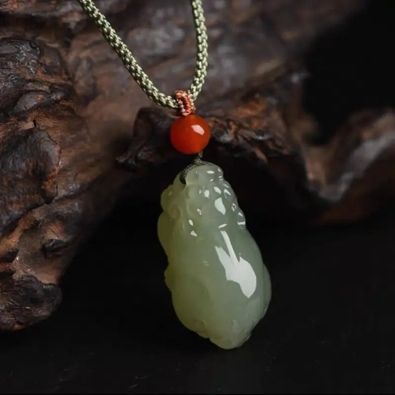 Hand-carved light green Hetian jade Pixiu pendant with a red agate bead on a green cord.