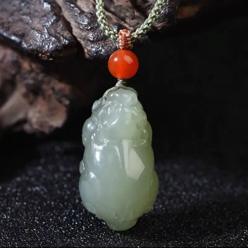 Front view of natural green Hetian jade Pixiu pendant with red bead accent on dark background.