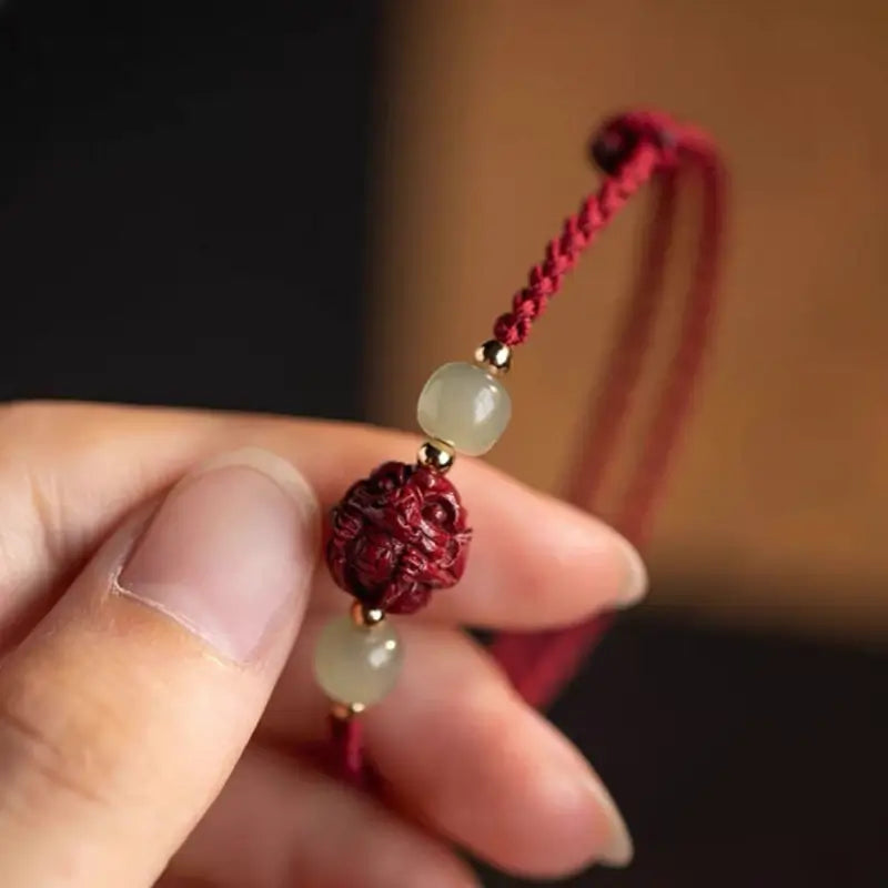 Hand holding red braided lucky bracelet showing intricately carved cinnabar zodiac bead and jade beads.