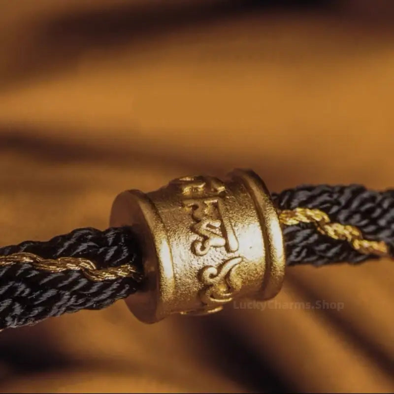 Macro detail of gold metallic cylindrical bead with carved Tibetan Six-Syllable Mantra script on braided cord.