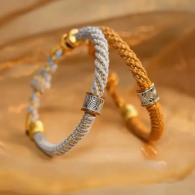 Pair of woven rope bracelets in light gray and yellow-gold with silver wave pattern charm beads.