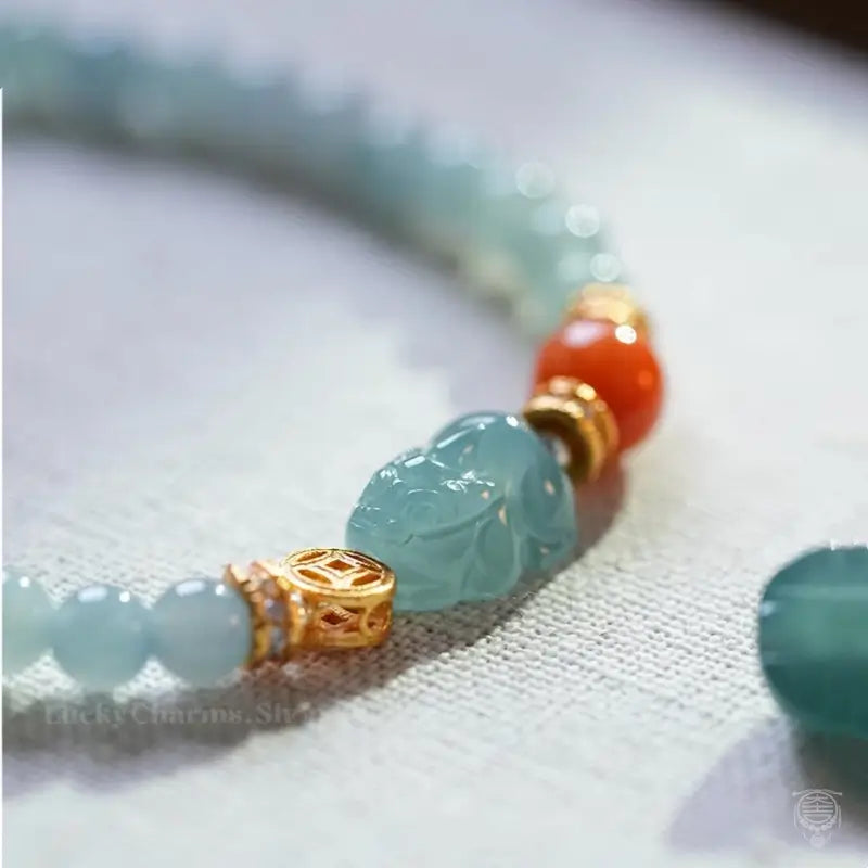 Macro detail of carved blue water jadeite Pixiu charm with gold coin spacer and red bead.