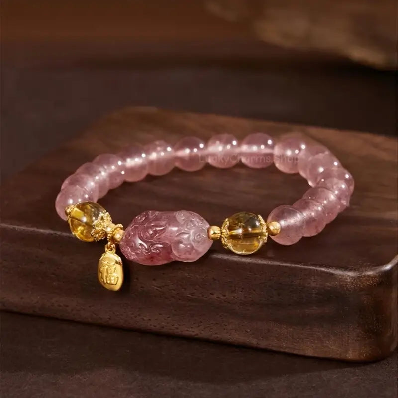 Palace Museum strawberry quartz Pixiu bracelet with yellow citrine beads and gold Fu charm on wooden block.
